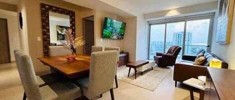 Una sala de estar moderna con comedor, un televisor montado en la pared y un amplio ventanal con vistas a la ciudad.