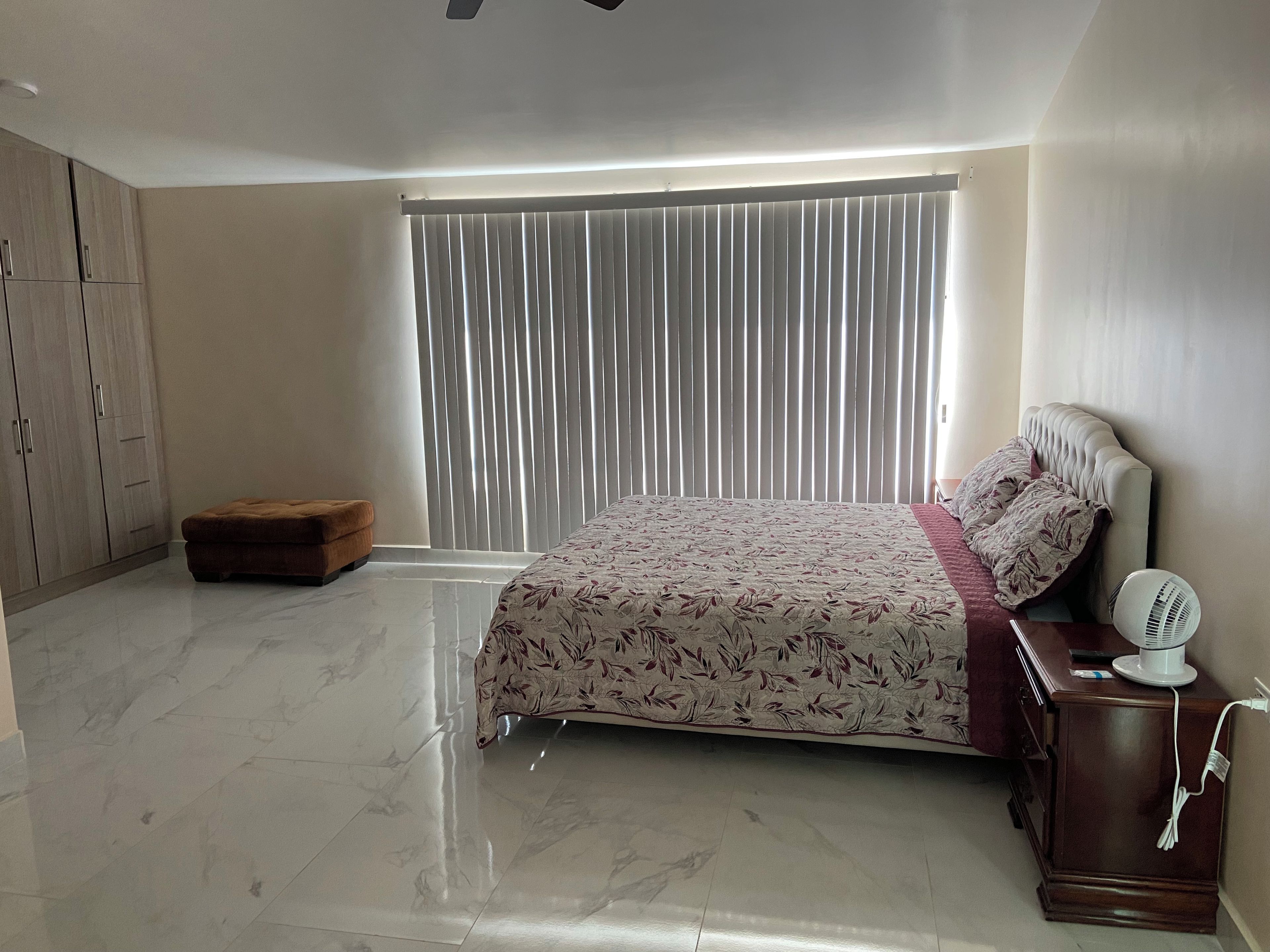 A bedroom with a bed, a bedside table with a fan, a wardrobe, and a bench.