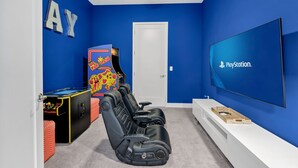 A room with a blue wall, a PlayStation TV, a gaming arcade machine, and a row of black gaming chairs. - Enormous 8 Bedroom Resort Home with View of 18th Hole near Disney with Resort &. (Kissimmee)