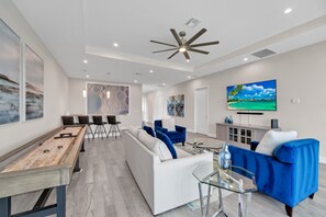 Un salon moderne avec une table de billard, des canapés bleus, une table basse en verre et un grand téléviseur à écran plat fixé au mur.