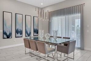 Une salle à manger moderne avec une table en verre, six chaises, des œuvres d’art abstraites accrochées au mur et une grande fenêtre avec des rideaux.