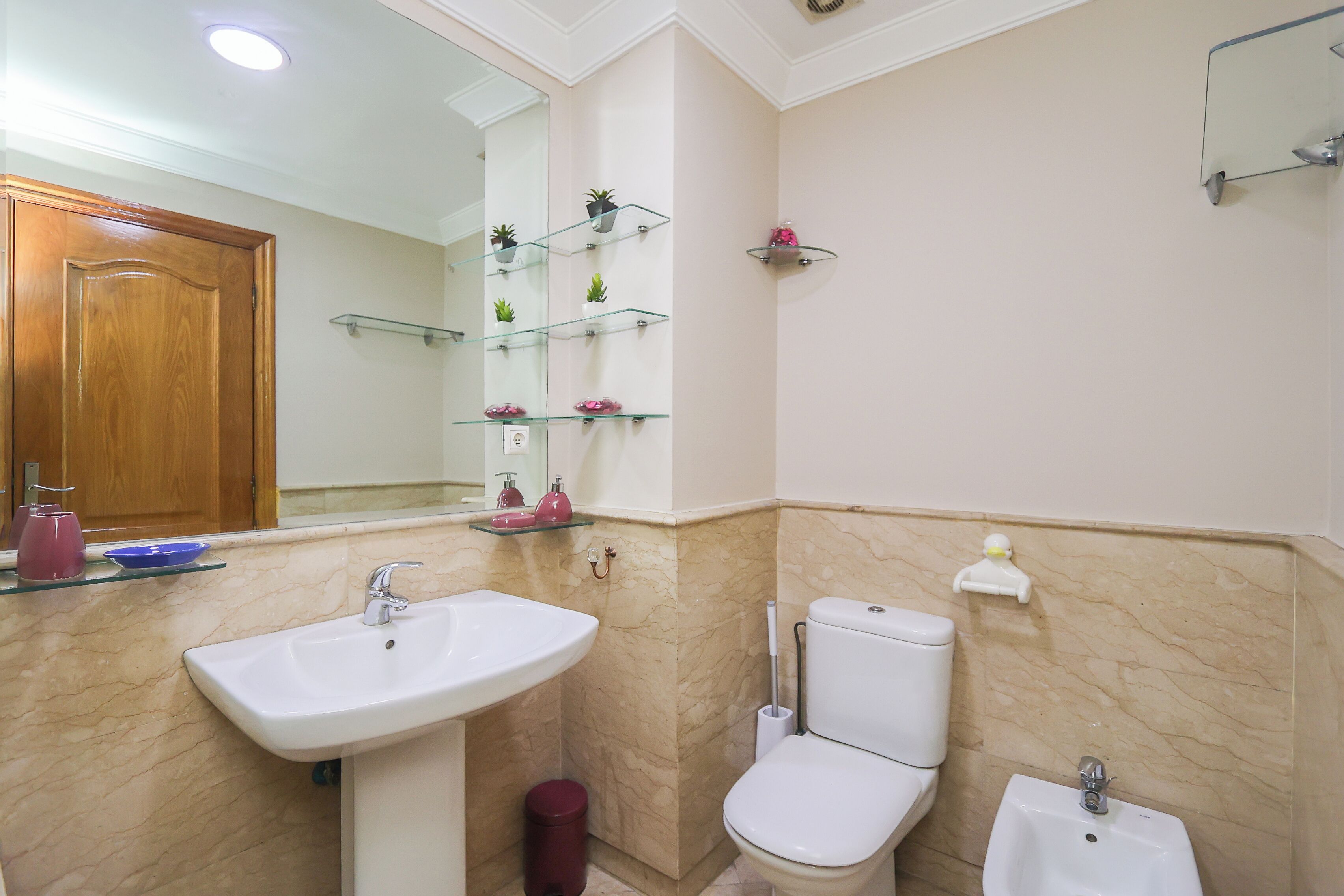 Un baño con lavamanos blanco, inodoro y bidé, paredes de mármol y puerta de madera.