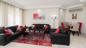 Uma sala de estar com um sofá de couro preto, almofadas vermelhas, um tapete vermelho estampado, um espaço de jantar com mesa e cadeiras, e obras de arte nas paredes.