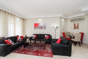 Ein Wohnzimmer mit einer schwarzen Ledersofa, roten Kissen, einem roten Teppich mit Muster, einem Essbereich mit Tisch und Stühlen sowie Kunstwerken an den Wänden.