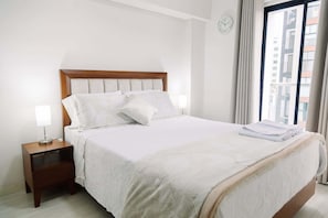 A neatly made bed with white linens, a wooden headboard, a bedside table with a lamp, and a view of buildings through a window. - Newly Renovated 2BR Apartment in Lima (Lince)