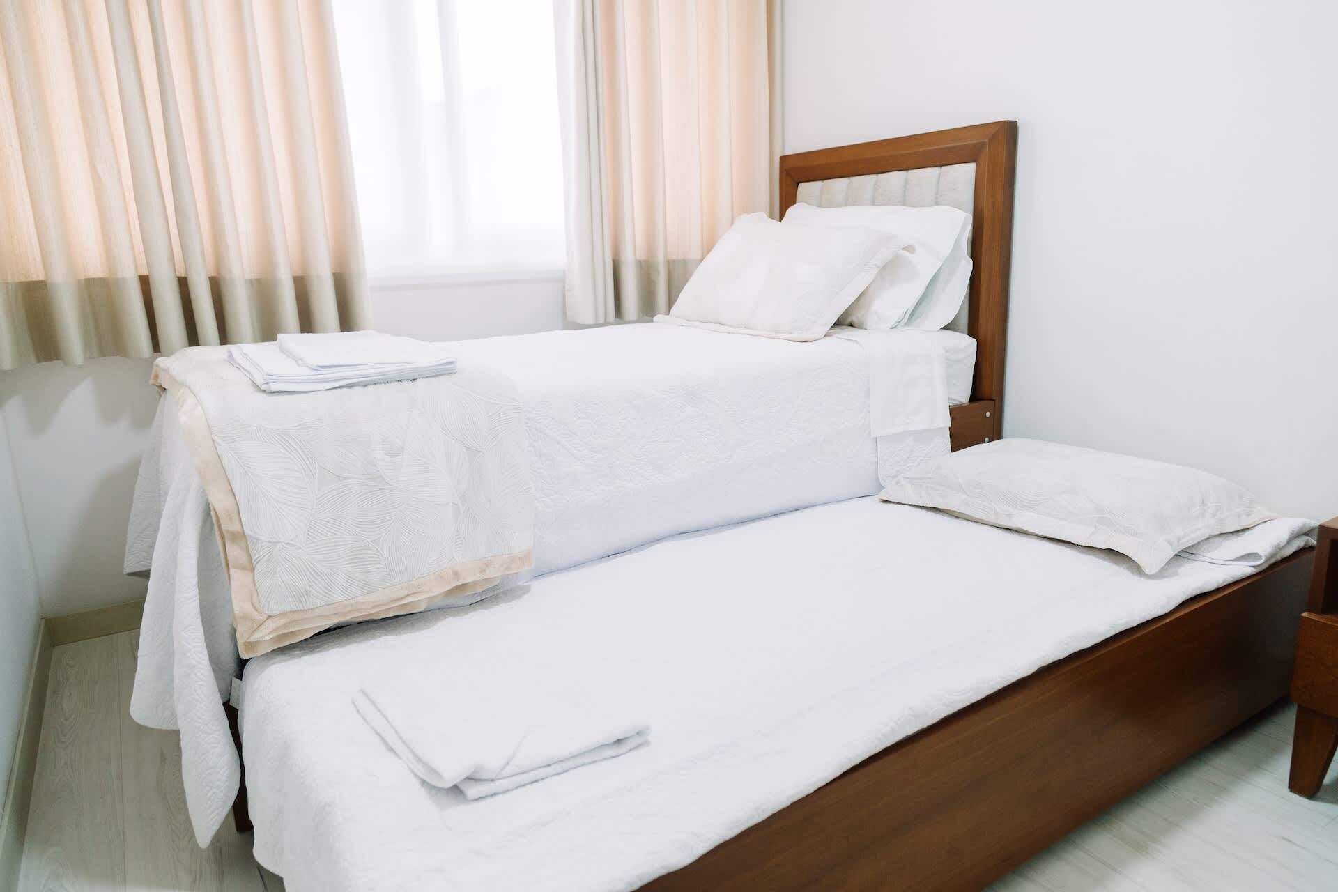 A neatly made bed with a wooden headboard and white linens in a room with light-colored walls and curtains.