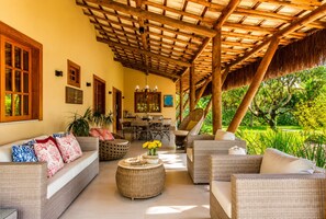 A covered outdoor seating area with wicker furniture, a dining table, and a view of a lush garden.