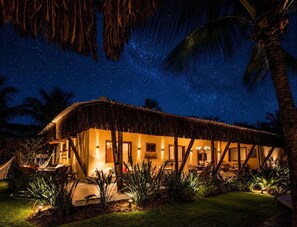 Un bâtiment à toit de chaume, doté de nombreuses fenêtres, entouré de plantes tropicales et de palmiers, sous un ciel nocturne étoilé.