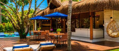 A tropical resort with a pool, wooden deck, lounge chairs, and a thatched-roof building.