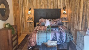 Quarto com teto de madeira, cama com roupa de cama estampada, duas luminárias de mesa, cômoda e radiador.