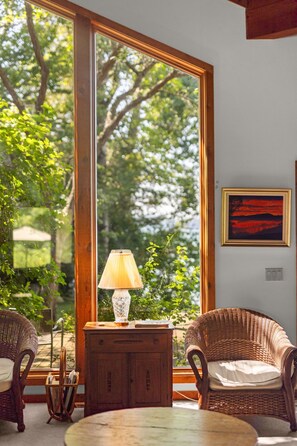 A room with a large window, wicker furniture, a table, and a lamp.