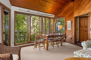 A dining area with a wooden table and chairs, large windows, and a view of trees.