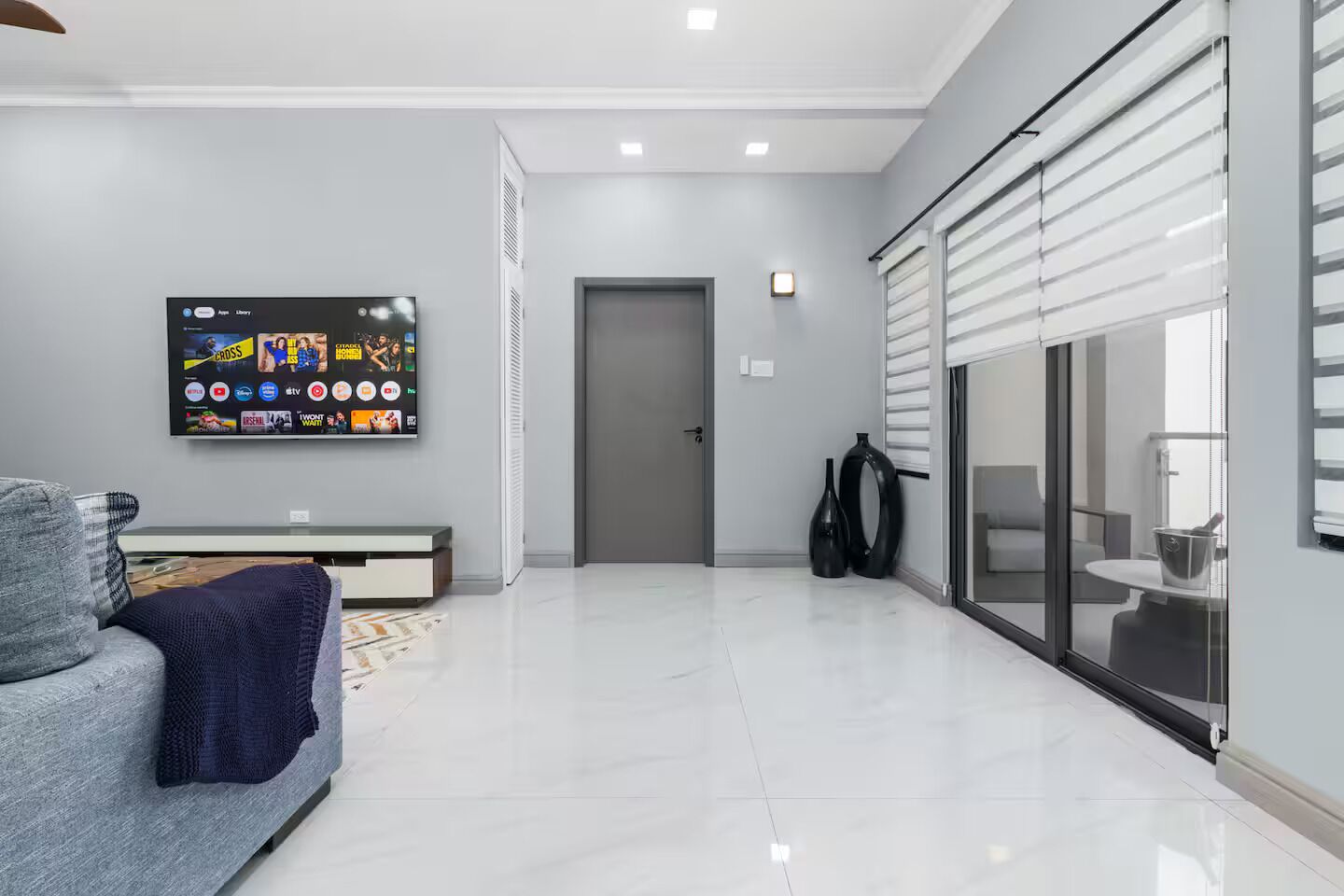 Uma sala de estar moderna com uma TV de tela plana na parede, um sofá cinza, uma mesa de centro e uma porta de vidro deslizante que dá para uma varanda.