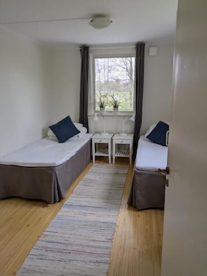 A room with two single beds, a small table, a chair, a window with curtains, and a ceiling light.