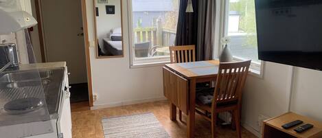A compact room with a kitchenette, a dining area with a table and chairs, a bed, a TV, and a window with curtains.