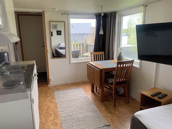 A compact room with a kitchenette, a dining area with a table and chairs, a bed, a TV, and a window with curtains.