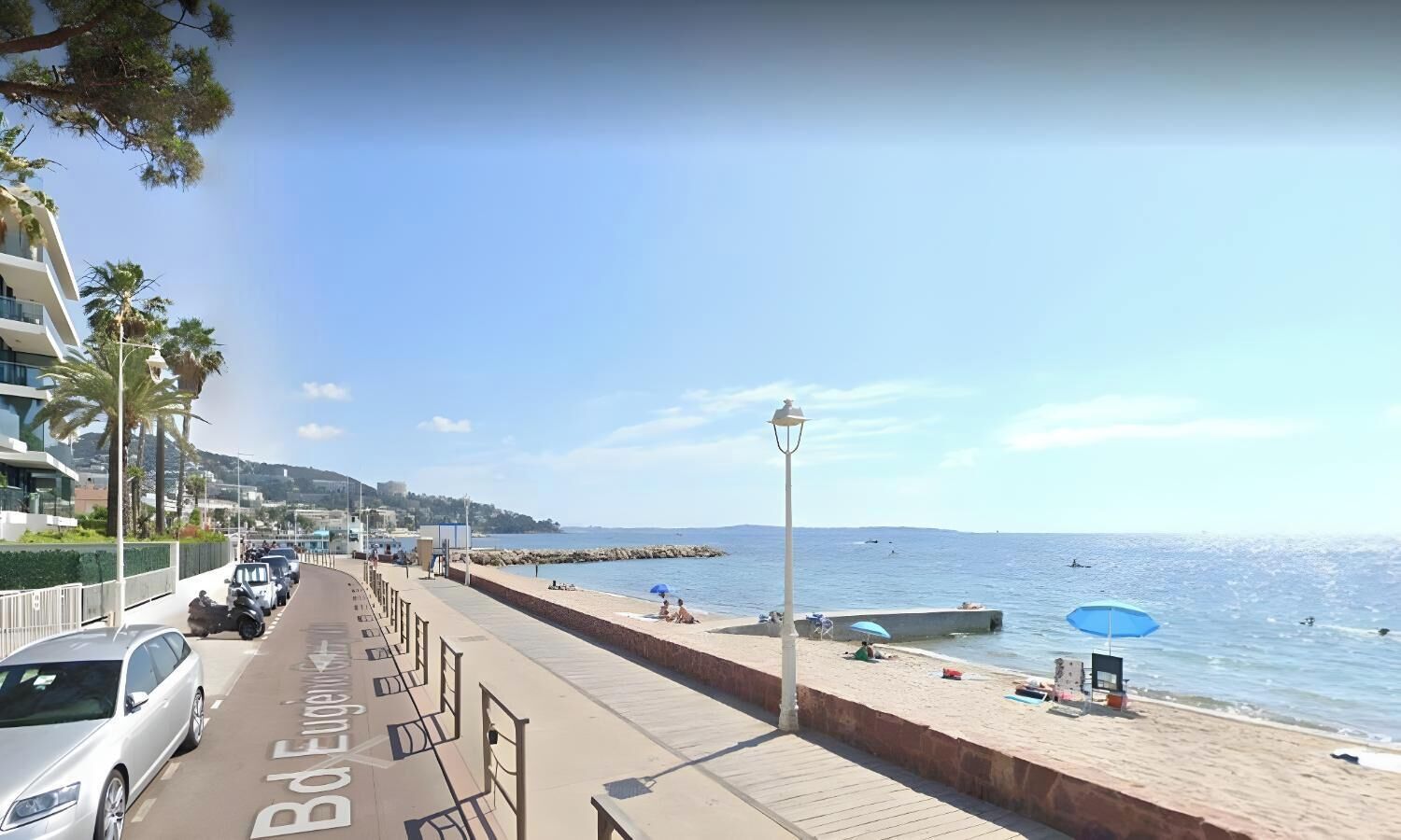 Une promenade en bord de mer, avec un chemin piétonnier pavé, des voitures garées et une vue sur la mer.