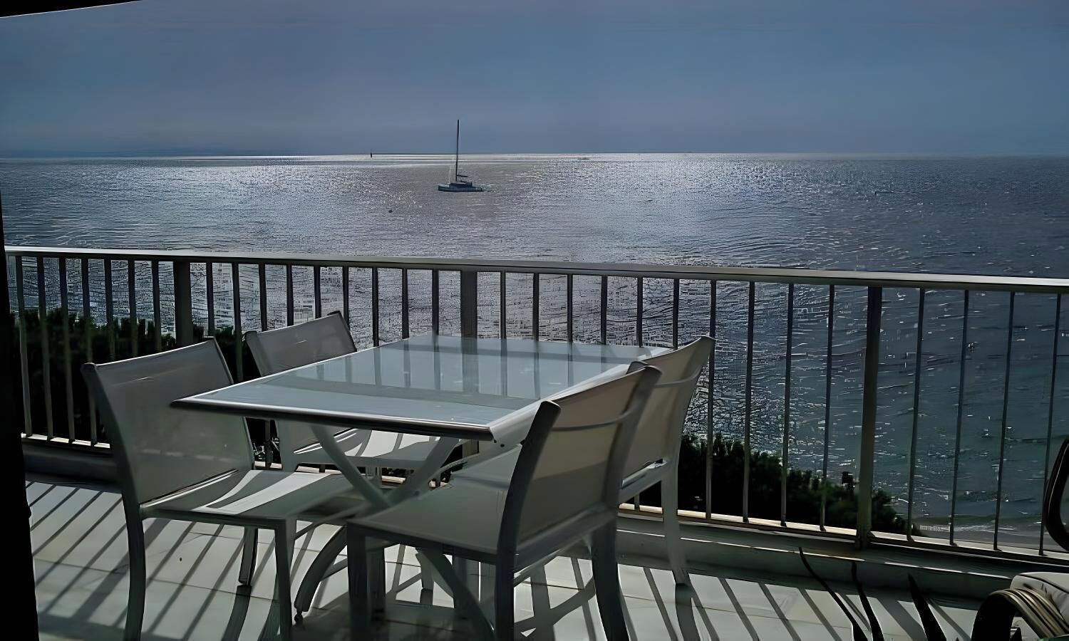 A balcony with a glass table and chairs, overlooking a calm sea with a sailboat in the distance.