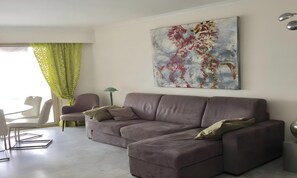 Ein Wohnzimmer mit einer Eckcouch, einem Esstisch mit Stühlen, einem grün gemusterten Vorhang und einem großen abstrakten Gemälde an der Wand.