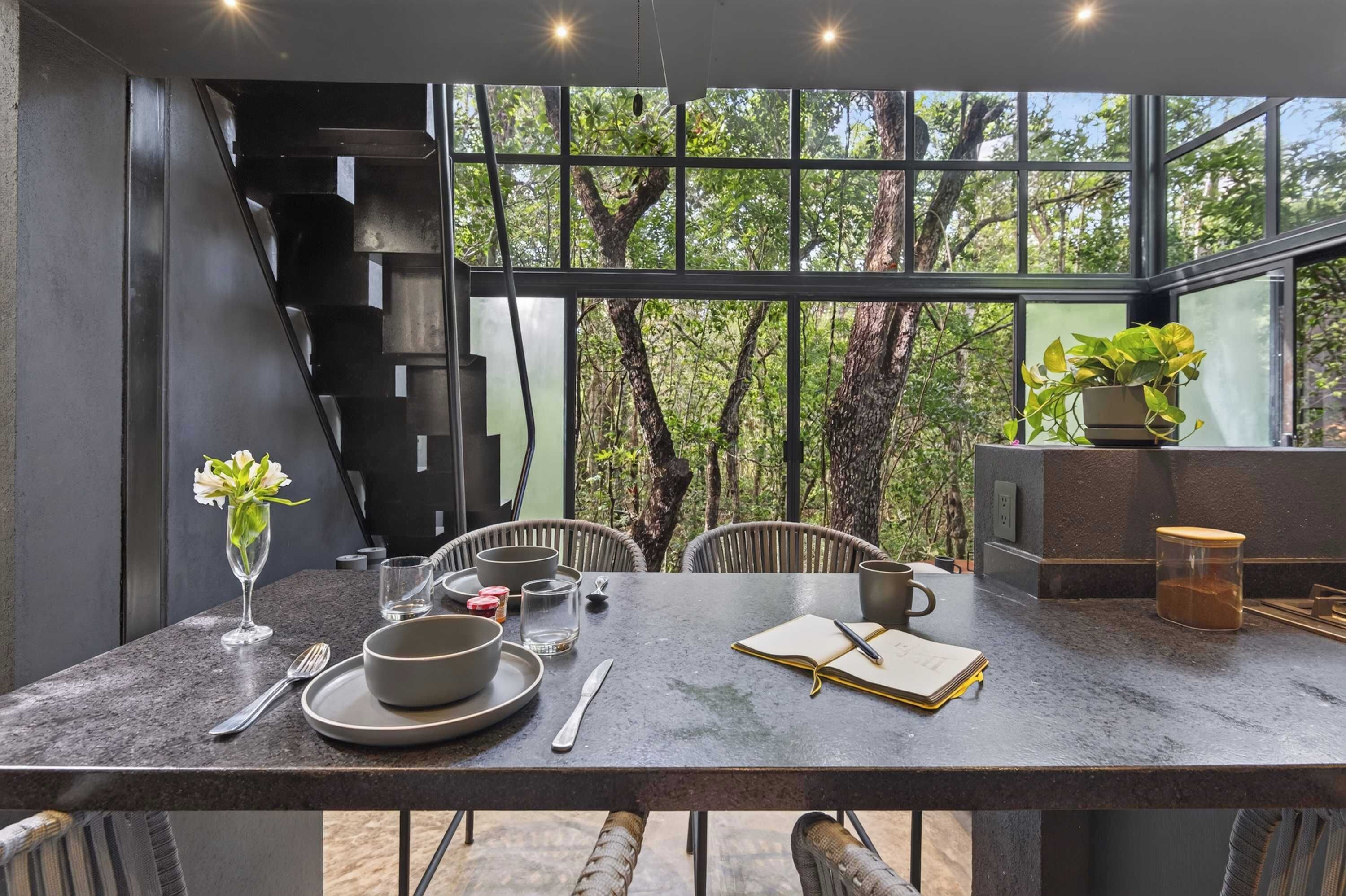 Un comedor moderno con una mesa dispuesta para dos personas, un florero y vistas a árboles a través de amplias ventanas.