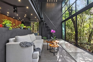 A modern interior with a sofa, a coffee table, and a staircase leading to a balcony with large windows overlooking a forest. - Tree60:· Jungle Villa Private Rooftop&Wood-Hot Tub (Chemuyil)