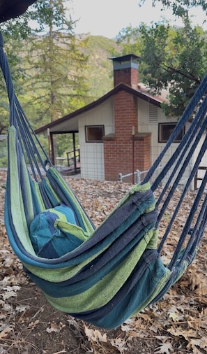 A hammock with a blue and green fabric, set against a backdrop of a building with a brick chimney and a white door.