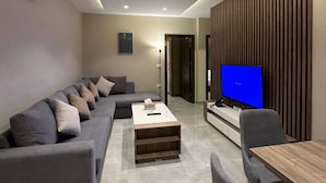 A modern living room with a sectional sofa, a coffee table, a TV, and a dining area. - 302 Al-najeh JRE Apartments For Rent ave (Amman)