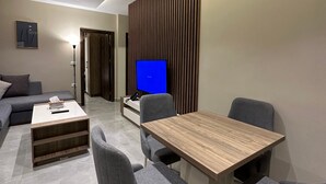 A modern living room with a wooden dining table, grey upholstered chairs, a flat-screen TV, and a sofa. - 302 Al-najeh JRE Apartments For Rent ave (Amman)