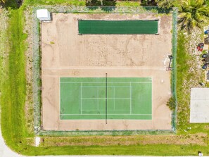 An aerial view of a tennis court surrounded by a fence, with a small building and parked vehicles nearby. - Adorable 2 Bedroom Condo - Pool & Prime Location (Fort Myers)