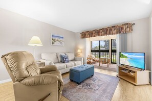 Een woonkamer met een bank, een poef, een televisie en uitzicht op de omgeving.