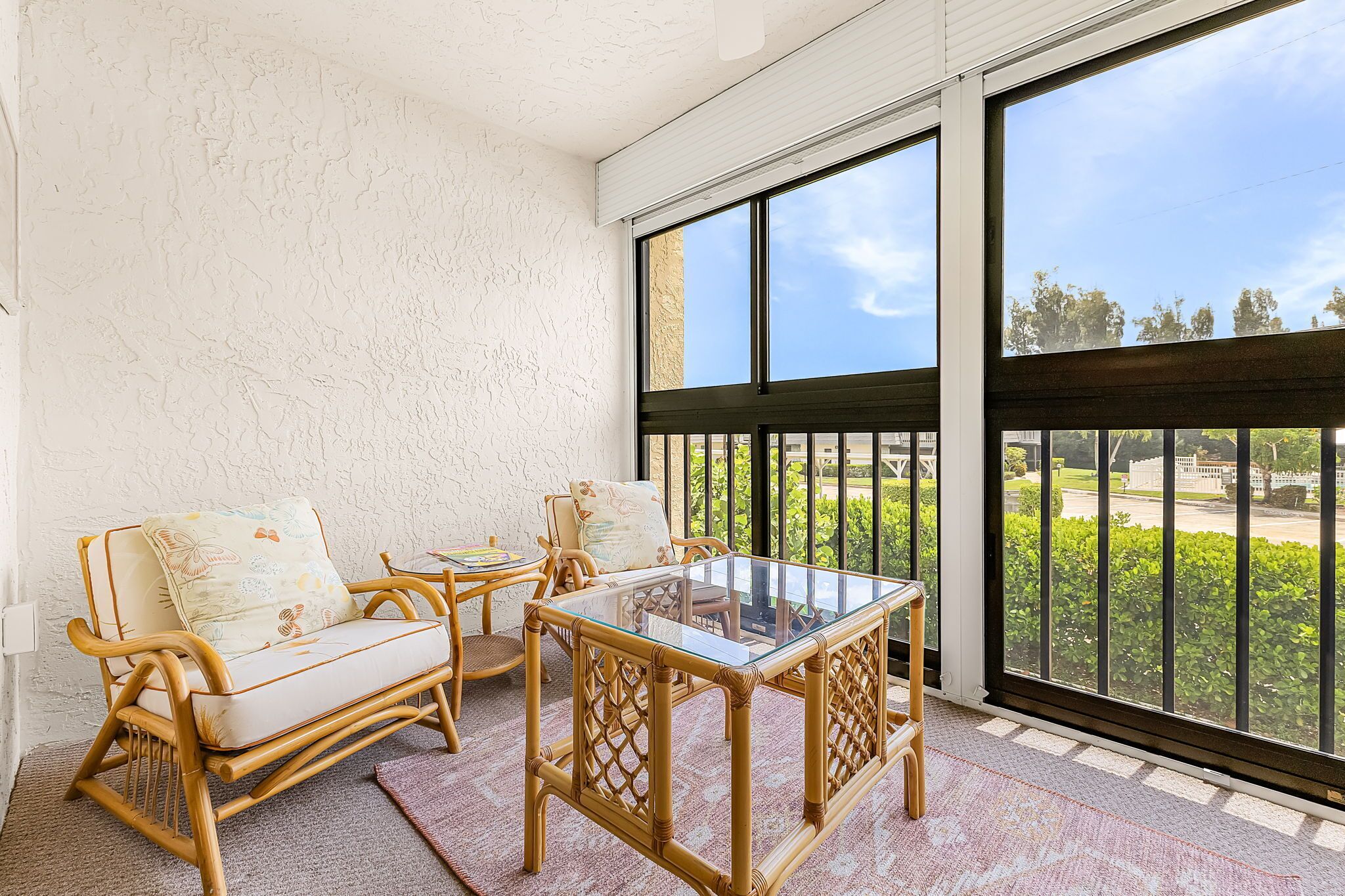 A balcony with wicker furniture, a glass table, and a view of greenery.
