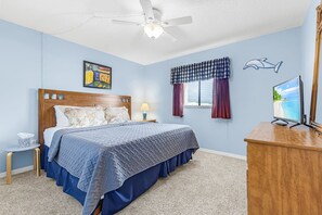 Een slaapkamer met een bed, een plafondventilator, een raam met gordijnen, een aan de muur bevestigde televisie en een klein tafeltje met een lamp.