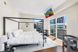 A modern bedroom with a four-poster bed, a city view balcony, and a large window.