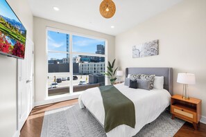 Un dormitorio moderno con un ventanal grande, una cama con muchas almohadas, una mesita de noche con una lámpara y un cuadro en la pared.