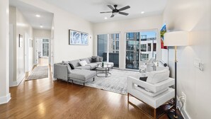 A spacious living room with a grey sectional sofa, a round coffee table, and a white armchair. There is a ceiling fan, a large window with a view of the outside, and a floor lamp. - Warm & Modern 3Bdrm NoLibs Condo - Sleeps 7! (Philadelphia)