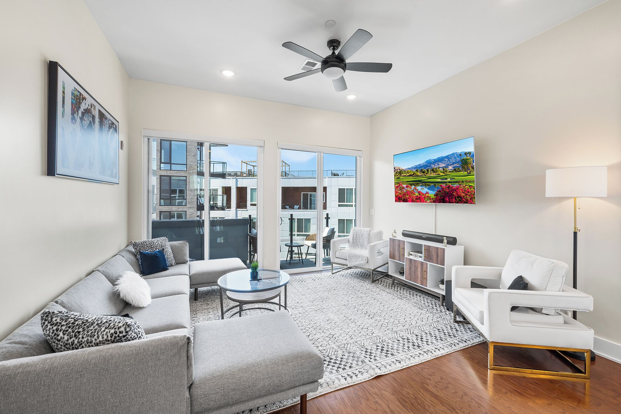A modern living room with a grey sofa, white armchair, and a coffee table. There is a ceiling fan, a painting, and a lamp.