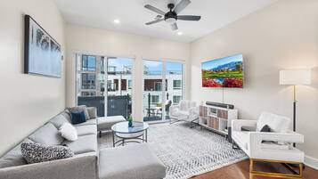 A modern living room with a grey sofa, white armchair, and a coffee table. There is a ceiling fan, a painting, and a lamp.