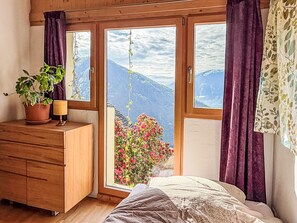 A bedroom with a wooden dresser, a bedside lamp, a bed, and a view of mountains through a sliding door. - Casa Serena by Interhome (Mergoscia)