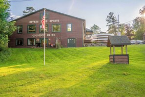 A riverside sports lodge with a grassy lawn, a flagpole, and a covered area with canoes. - Rangeley Lakefront 3BR | Private Dock, Dogs OK (Rangeley)