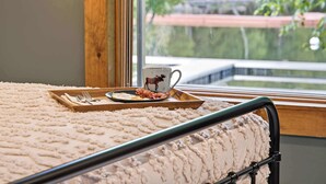 A bed with a fluffy white blanket, a tray with breakfast including eggs, bacon, and a mug with a moose design, and a window with a view of greenery and a bridge.