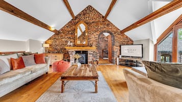 A loft-style living room with exposed wooden beams, a brick fireplace, and a television.