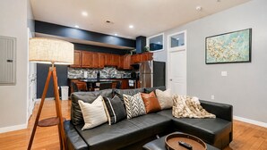 A modern living room with a black leather sofa, a standing lamp, and a remote control on a wooden tray. - The Camac Hideaway - 2BR near Convention Center (Philadelphia)