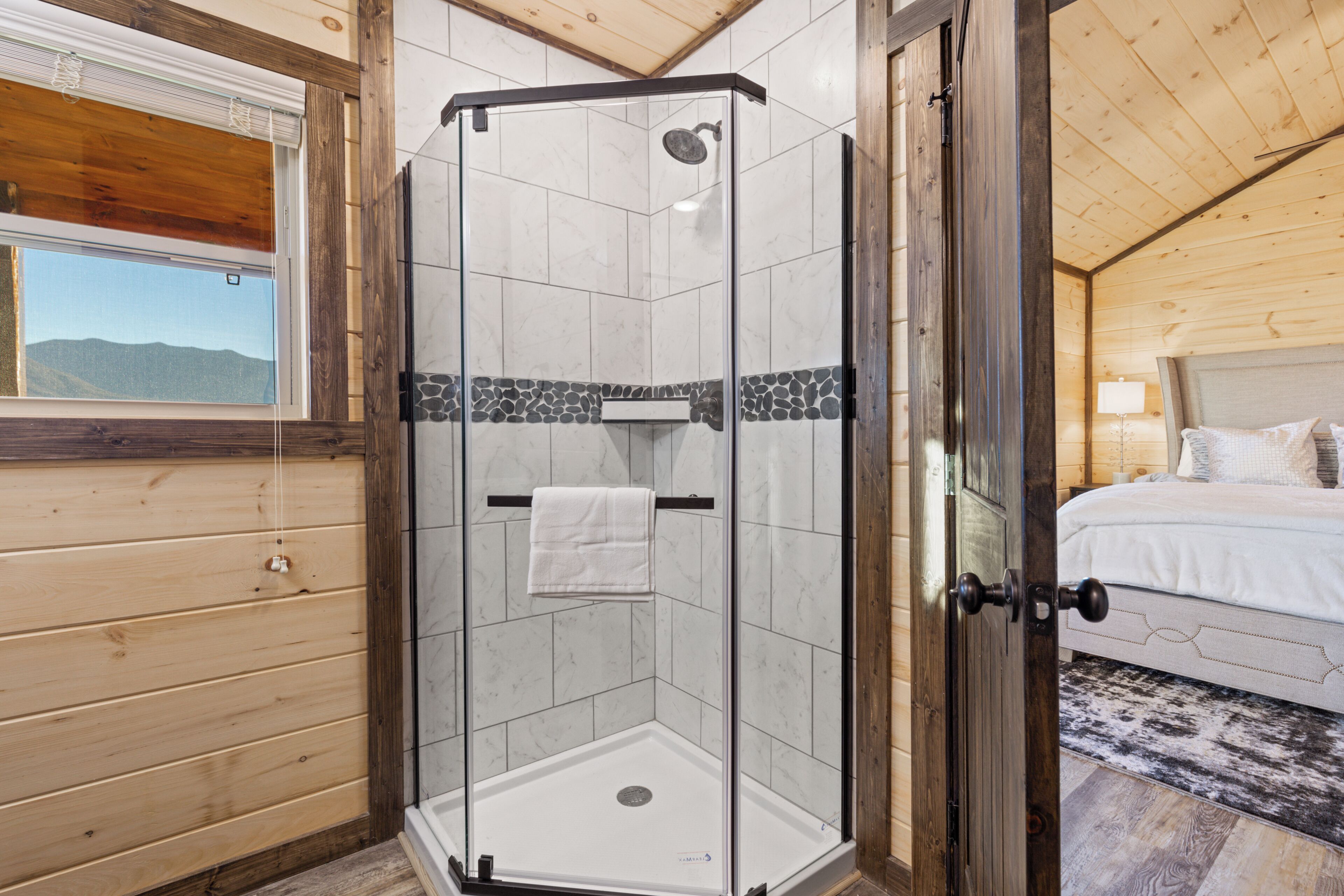 A glass-enclosed shower with a white towel hanging on the door handle, a wooden ceiling, and a bed with pillows in the adjacent room.