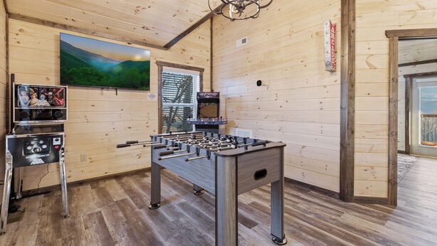 A wooden-floored room with a foosball table, a television, and a window with a view of a building. - The Overlook at Settlers Ridge (Sevierville)