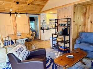 Ein gemütliches Wohnzimmer mit einer Ledersofa, einem Couchtisch und einem blauen Sofa. Es gibt einen Holzesstisch mit Stühlen und eine Küchenzeile mit einer Kaffeemaschine und Regalen.