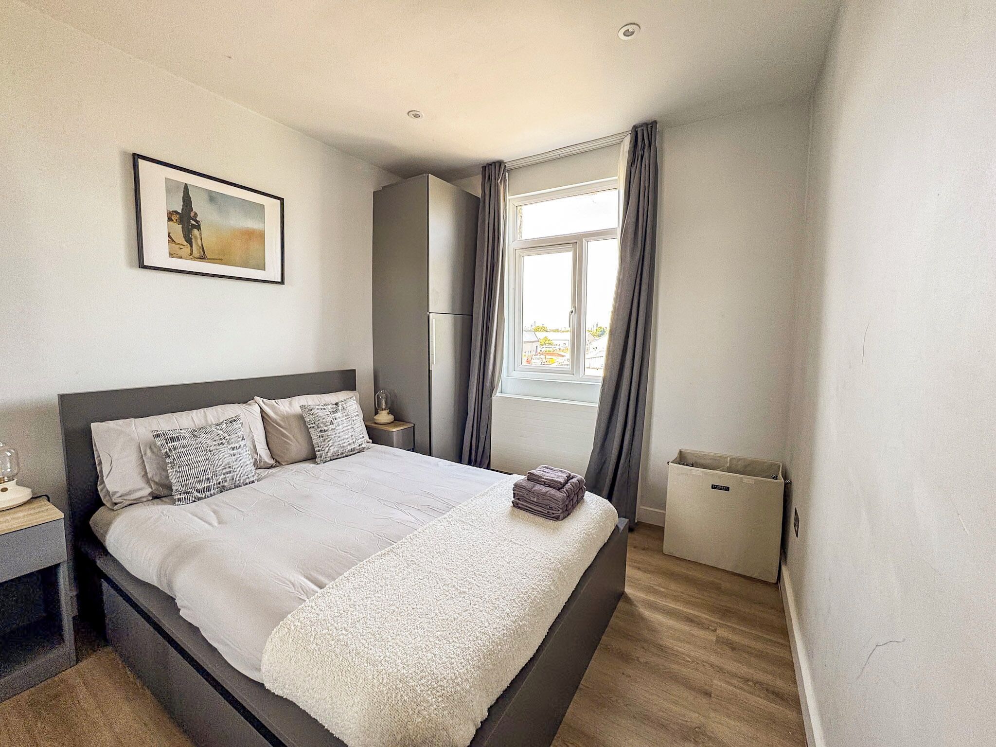 A bedroom with a bed, a nightstand, a window with curtains, a radiator, and a framed picture on the wall.