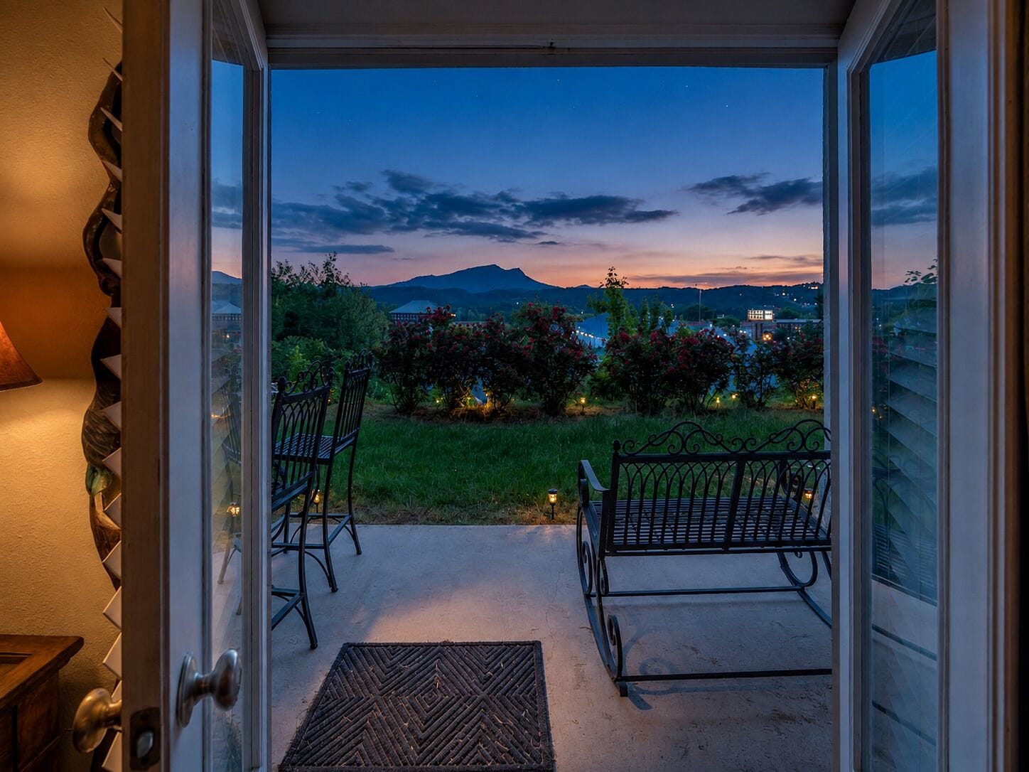 Une porte ouverte donne sur un balcon où se trouvent deux chaises et une petite table. Le balcon offre une vue sur un jardin, avec en arrière-plan des montagnes lointaines et un paysage urbain au crépuscule.