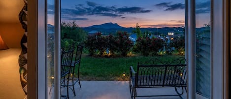 An open door leading to a balcony with two chairs and a small table, overlooking a garden with a view of distant mountains and a cityscape at dusk.