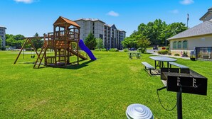 Ein Spielplatz mit einer Holzspielkonstruktion, einer blauen Rutsche, Picknicktischen und einem Grill in einer grünen Rasenfläche.