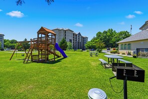 Ein Spielplatz mit einer Holzspielkonstruktion, einer blauen Rutsche, Picknicktischen und einem Grill in einer grünen Rasenfläche.